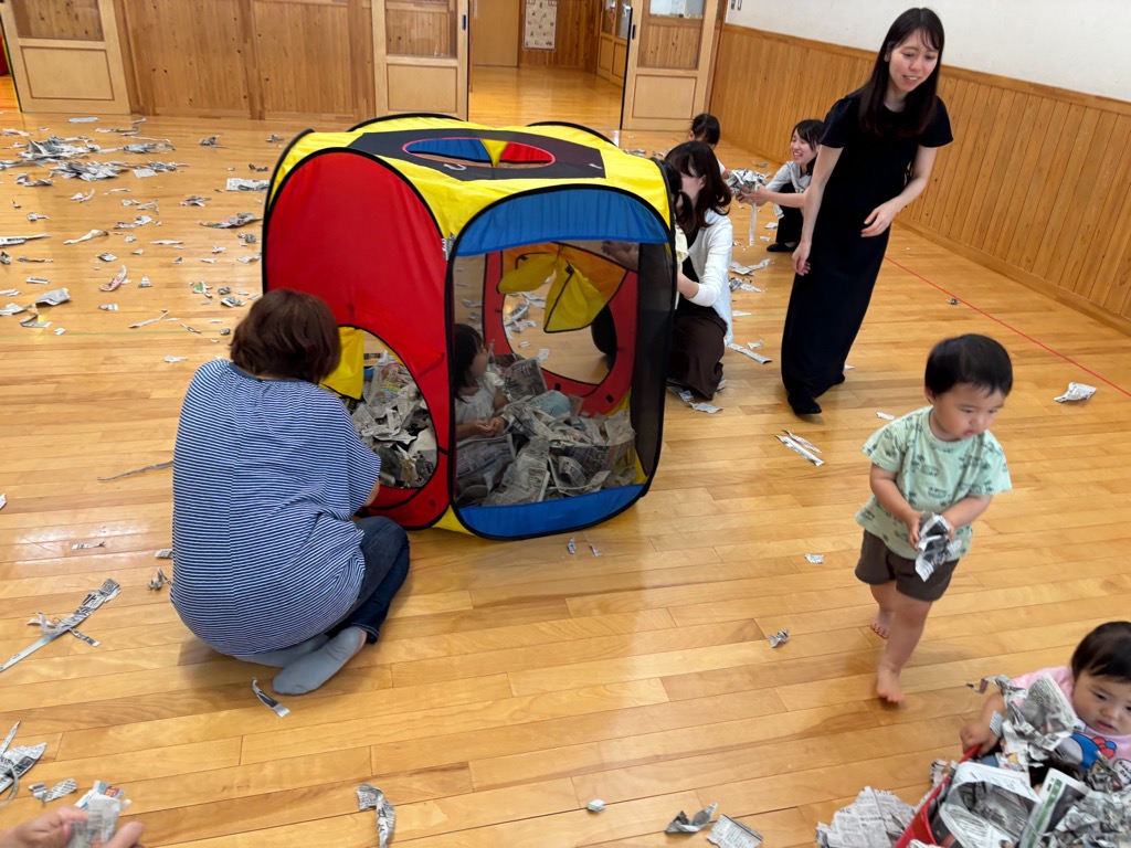 こあらの日の新聞紙あそび 新聞紙をいっぱい持って友達の箱車に乗せようとしているところ