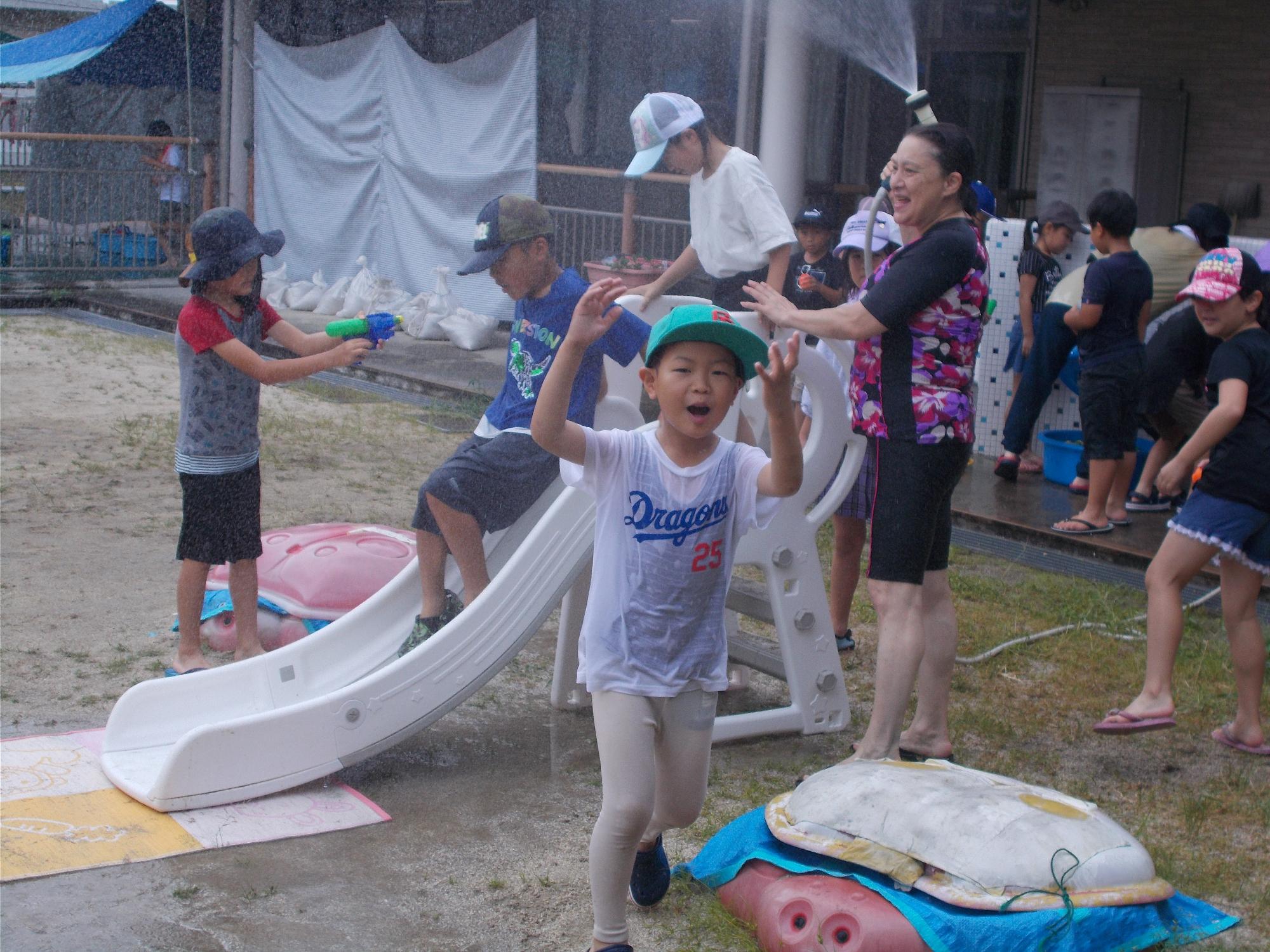 水遊びで両手を挙げて大喜びしている男の子。その後ろではおもちゃの滑り台でウォーター滑り台を楽しんでいる子どもたち