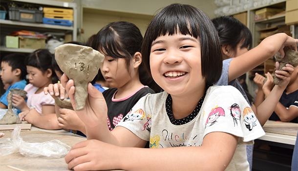 完成した製塩土器を片手に笑顔を見せる女の子、後ろには真剣なまなざしで粘土をさわり製塩土器を形成している児童たちが見える