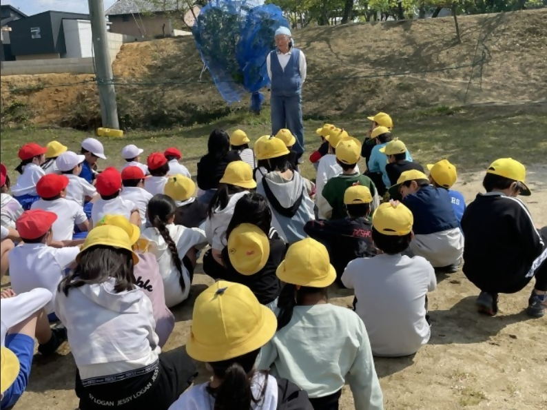 学校の南側の果樹園で地域の方から果物の育て方を熱心に聴く子どもたち